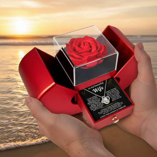 A red heart-shaped gift box on a white background, opened to display a preserved red rose in a clear acrylic case and a red jewelry box drawer with a necklace and printed message for a wife.
